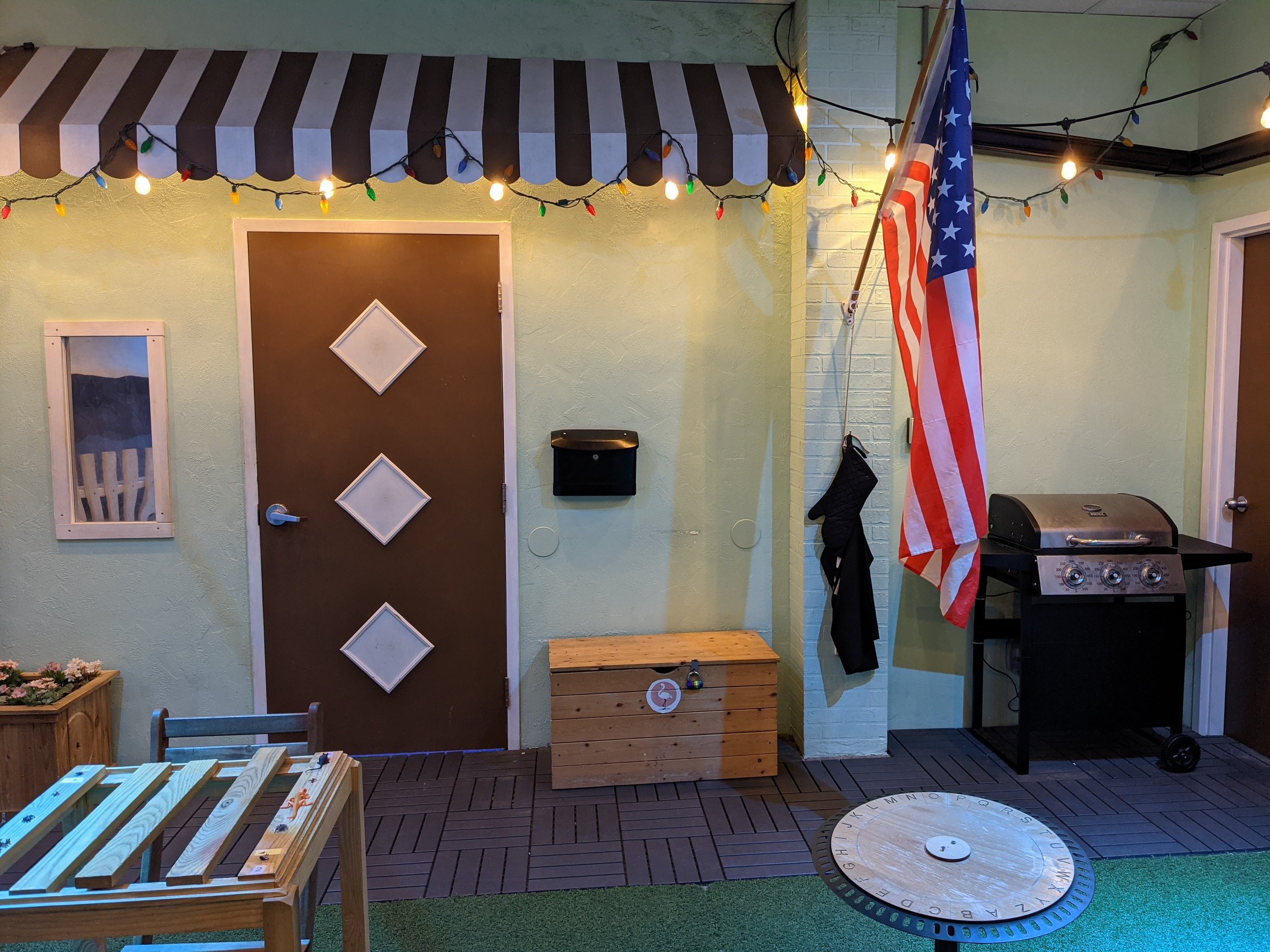 Outdoor patio with string lights, a picnic table, a wooden storage box, a barbecue grill, and American flag, decorated for a celebration.